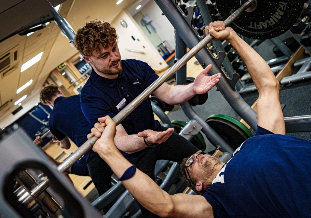 Personal trainer at health club in Woking assisting a man with a bench press, ensuring proper form and safety during a gym Woking strength training session