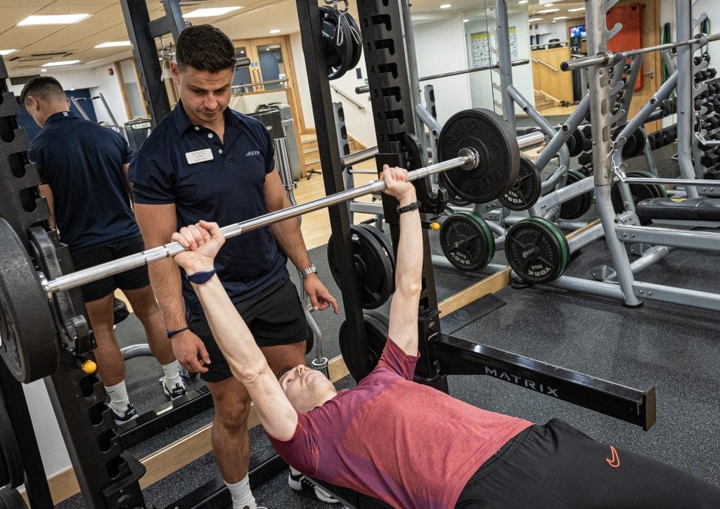 Personal trainer assisting client in bench press at gym in Woking