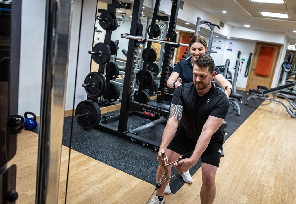 Female personal trainer assisting man in cable machine exercise at gym in Woking